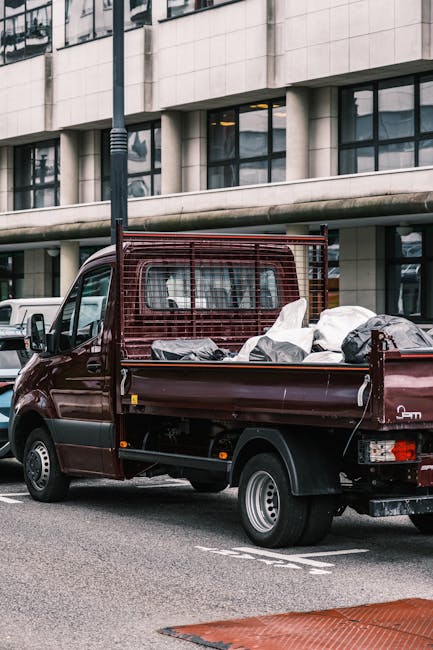 A maroon flatbed utility truck parked on an urban street in front of a modern building with large glass windows and an off-white facade. The truck's open cargo area is filled with various household ru
