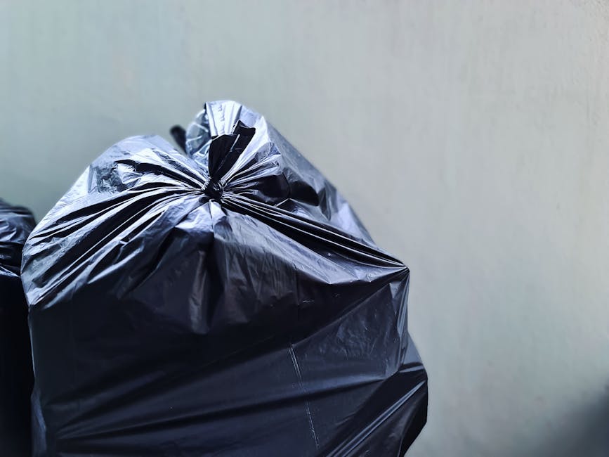 A large green industrial rubbish skip placed on a paved area adjacent to a dark painted brick wall. The skip is filled with black and red bags of building or garden waste, some of which are stacked on