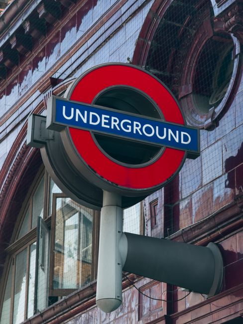 A low-angle view of a busy underground London tube station, showing the curved tiled ceiling with panels and a central lighting strip, as well as the platform with tactile yellow safety lines running